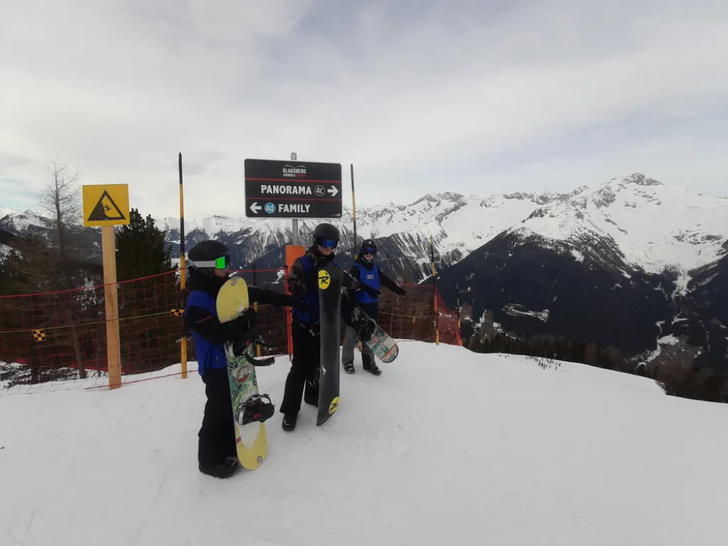 Zwei Snowboarder stehen mit ihren Brettern an einem Wegweiser vor Bergpanorama.